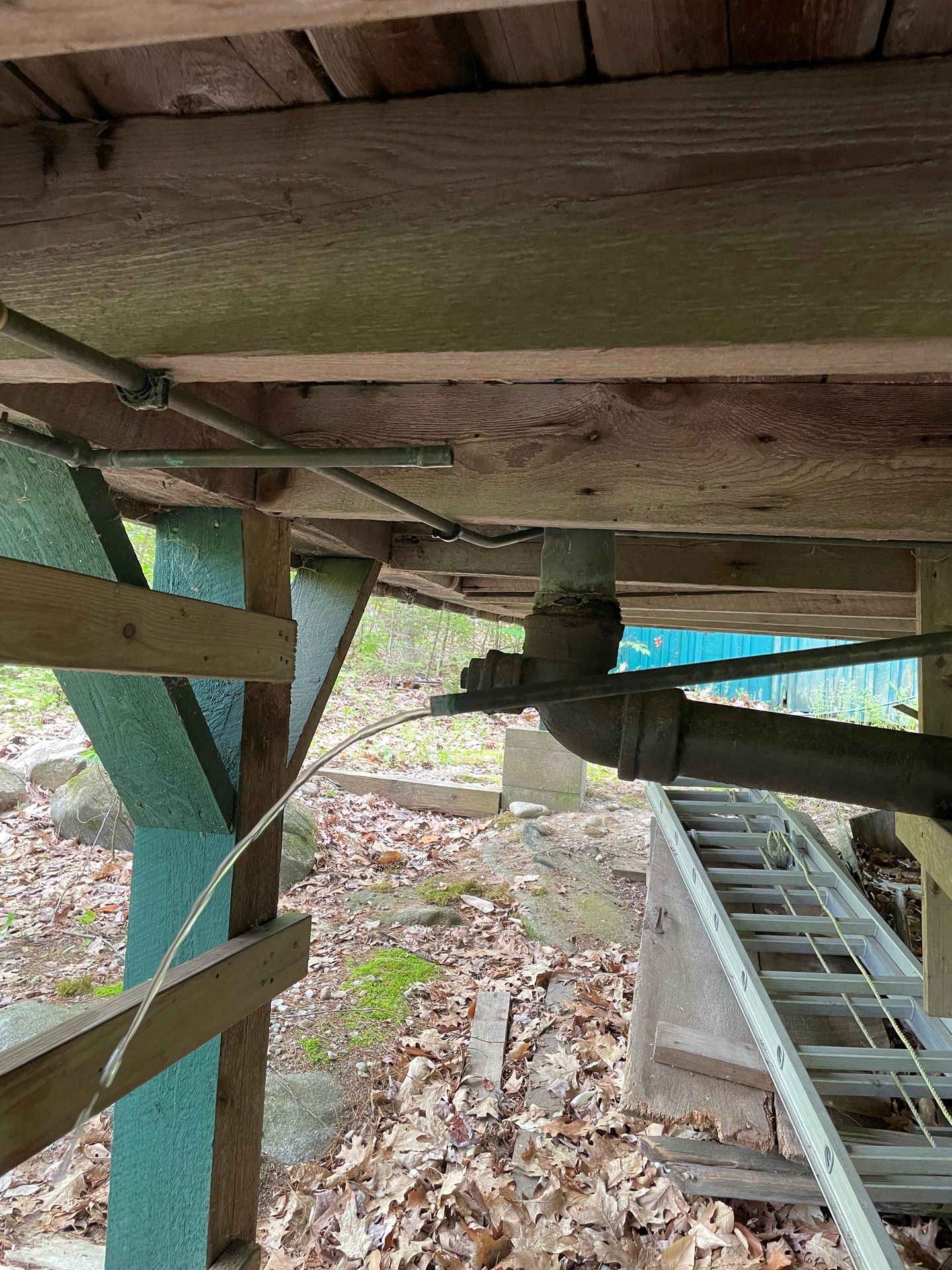 Underneath a wooden structure with pipes, wires, and scattered leaves on the ground.